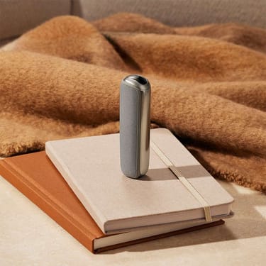Dispositivo Ploom con Cover Posteriore Lunar Silver in piedi sopra una pila di quaderni